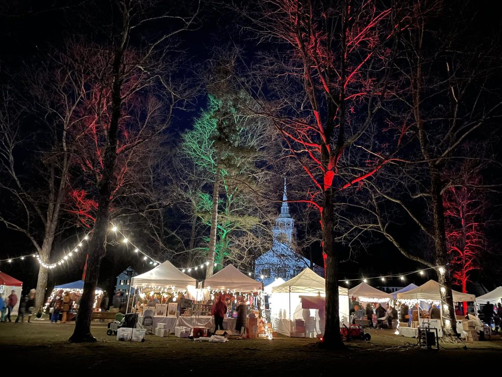 Nighttime scene of the Amherst German Christmas Market on the Village Green, showing white vendor tents, crowds browsing stalls, and trees illuminated with red and green lights, with the historic church steeple lit in the background.