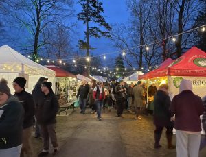 Close-up view of the Amherst German Christmas Market showing crowds of people walking between vendor stalls and food tents, with string lights strung overhead at dusk.